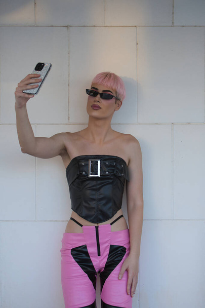 Привлекательный молодой гей, сильно накрашенный, с розовыми волосами, солнцезащитными очками, кожаным топом и брюками, фотографирующий на мобильный телефон, рядом со стеной из белых каменных блоков. LGTBIQ+. Вертикальное положение. - Фото, изображение