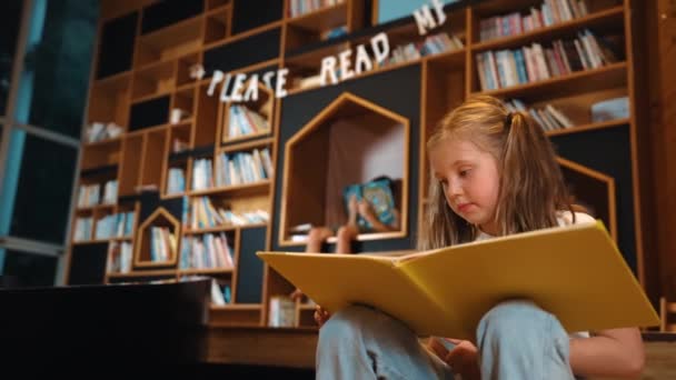 Joven chica caucásica inteligente recogiendo la lectura de un libro mientras está sentado en la biblioteca. Un niño inteligente aprendiendo, estudiando, abriendo libros en la biblioteca. Atractivo niño pasando página con fondo borroso. Erudición. - Imágenes, Vídeo