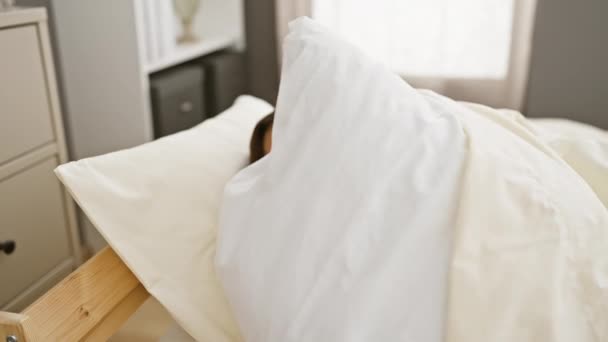 Mujer joven envuelta en un edredón blanco sonríe suavemente mientras descansa en un dormitorio con una decoración moderna. - Imágenes, Vídeo