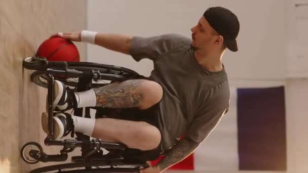 Függőleges, teljes hosszúságú portré fiatal, fogyatékossággal élő kaukázusi férfiról, aki kosárlabdázik kerekesszékben a beltéri pályán és kamerába néz - Felvétel, videó