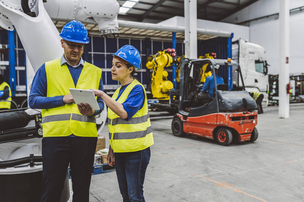 Two engineers in safety gear reviewing plans on a tablet in an industrial factory, forklift in background. - Photo, Image