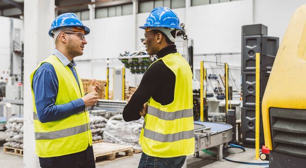 Dos ingenieros con sombreros duros y chalecos de alta visibilidad participaron en una discusión dentro de una instalación industrial. - Foto, imagen