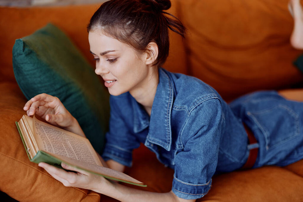 Γυναίκα χαλαρώνοντας στον καναπέ στο σπίτι, διαβάζοντας το βιβλίο σε denim πουκάμισο, άνετο και άνετο εσωτερικό σκηνικό - Φωτογραφία, εικόνα