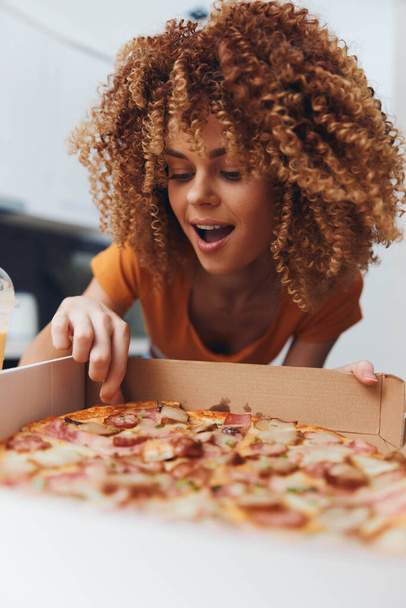 Mujer con el pelo rizado mirando una pizza recién horneada en una caja en la cocina - Foto, imagen