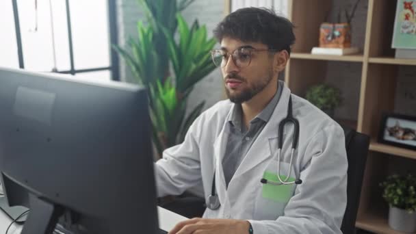 Arabischer Arzt mit Brille und Laborkittel arbeitet am Computer und telefoniert in einem Klinikbüro. - Filmmaterial, Video
