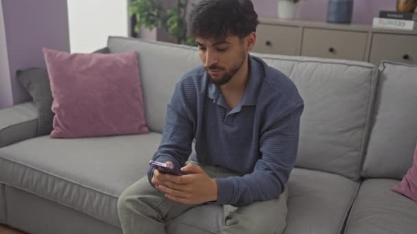 Jovem árabe homem, confiante e auto-confiante, imerso em pensamento profundo enquanto sentado em um sofá em casa, seu smartphone na mão e expressão sóbrio - Filmagem, Vídeo