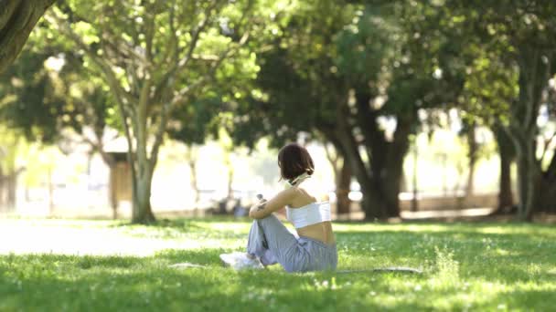Una mujer está sentada en el césped de un parque. Lleva una camiseta blanca y pantalones grises. - Metraje, vídeo