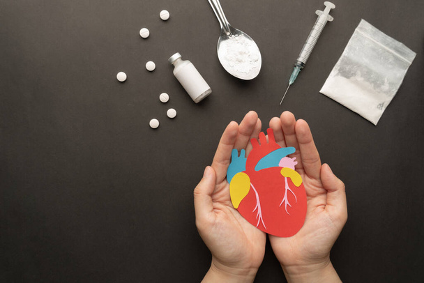 Επιδράσεις των φαρμάκων στην ανθρώπινη καρδιά. Χέρια κρατώντας κομμένο χαρτί από την καρδιά με σκόνη φαρμάκων, σύριγγα και χάπια σε σκούρο φόντο. Διεθνής ημέρα κατά της χρήσης ναρκωτικών. - Φωτογραφία, εικόνα