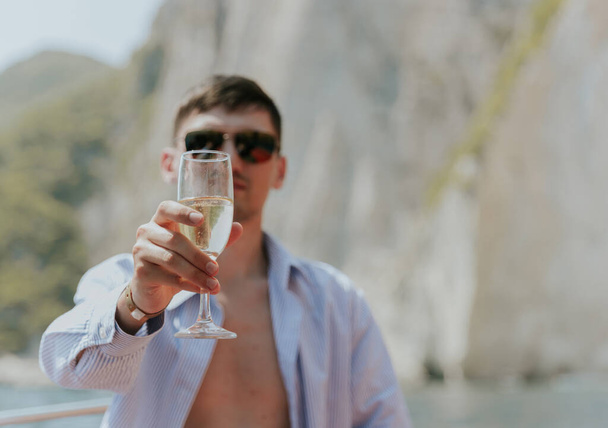 Portrait of one young Caucasian handsome guy in sunglasses sits at the stern of a boat, holds out a glass of champagne, looks into the camera and enjoys sailing on the sea on a private boat on a sunny - Photo, Image