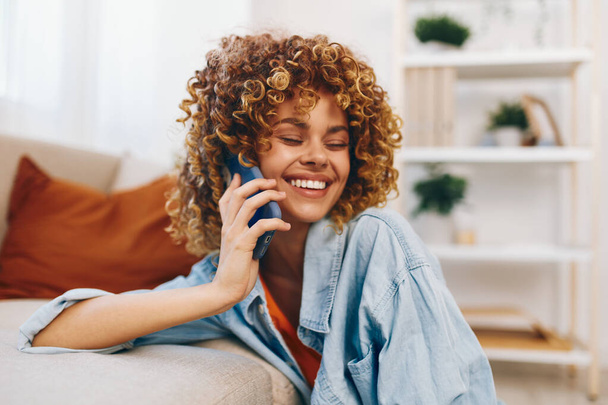 Wesoła kobieta w domu, siedzi na przytulnej kanapie z telefonem komórkowym, uśmiechnięta i ciesząca się grami online - Zdjęcie, obraz