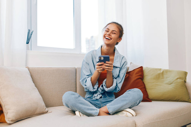 Accogliente soggiorno: una donna felice che tiene un telefono cellulare su un divano a casa - Foto, immagini