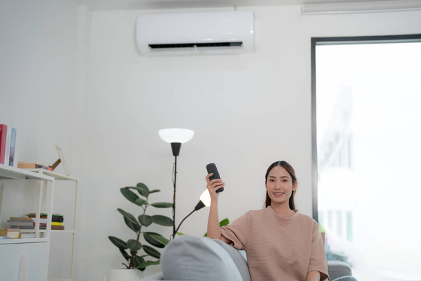Jeune femme souriante utilisant son smartphone dans un salon bien éclairé pour contrôler à distance un climatiseur mural élégant, illustrant la technologie de la maison intelligente et la commodité moderne - Photo, image