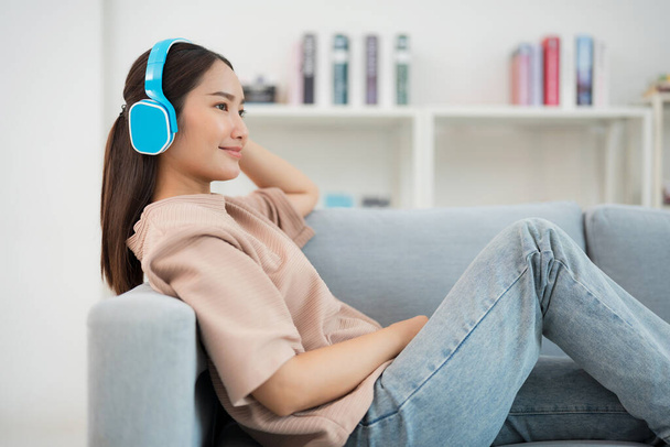 Mujer joven sonriente se relaja en un sofá con los ojos cerrados, disfrutando de su música favorita en auriculares azules en una acogedora sala de estar con una estantería en el fondo - Foto, Imagen