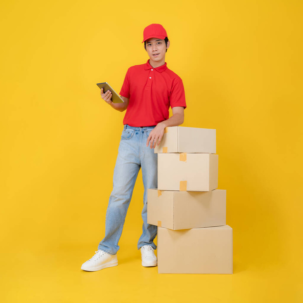 Εύθυμη παράδοση άνθρωπος σε ένα κόκκινο πόλο και καπάκι στέκεται δίπλα σε μια στοίβα των πακέτων, κρατώντας ένα ψηφιακό δισκίο σε ένα ζωντανό κίτρινο φόντο, που απεικονίζει την αποτελεσματικότητα και online διαχείριση της παραγγελίας - Φωτογραφία, εικόνα