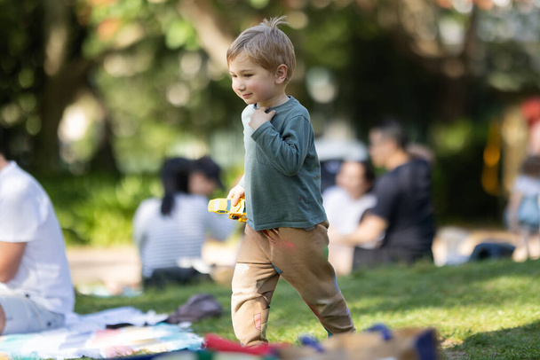 Un ragazzino sta giocando con una macchinina in un parco. Ci sono altre persone sullo sfondo, alcune delle quali sono sedute su coperte. La scena è vivace e giocosa - Foto, immagini