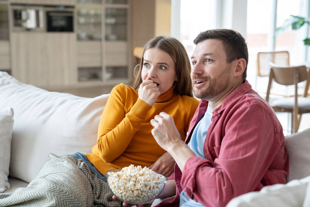 Surprised amazed man and woman watching TV movie together sitting on couch at home eating popcorn. Family couple wife husband hooked on series. Pastime, leisure, watch television rest relax weekend. - Photo, Image