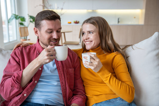 Ευτυχισμένο ζευγάρι άντρας και γυναίκα πίνοντας τσάι καφέ κάθεται στον καναπέ στο σπίτι, απολαμβάνοντας μιλώντας τρυφερές αγκαλιές. Οι αγαπημένοι σύζυγοι παντρεύονται και κάνουν ωραία συζήτηση. Αρμονική σχέση γάμου - Φωτογραφία, εικόνα