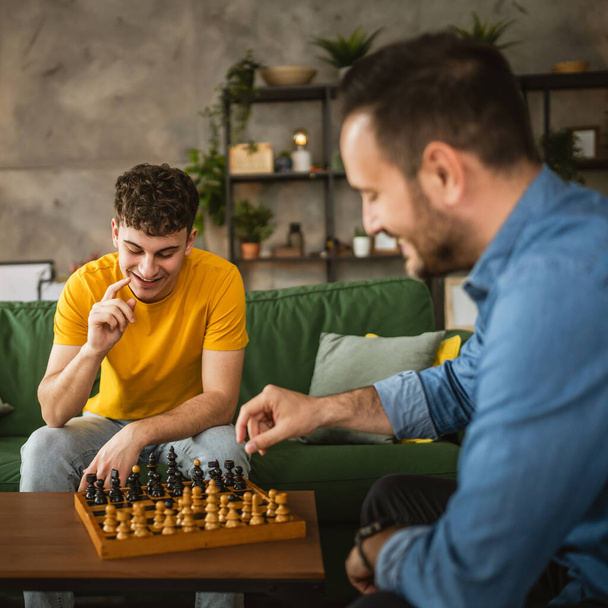 młody i dorosły biały mężczyzna przyjaciele, kuzyni lub bracia grać w szachy razem w domu - Zdjęcie, obraz