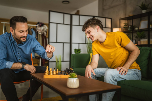 νέοι και ενήλικες καυκάσιοι φίλοι, ξαδέλφια ή αδελφοί παίζουν σκάκι μαζί στο σπίτι - Φωτογραφία, εικόνα
