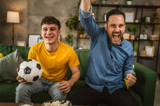 Mladý kavkazský a dospělý kavkazský muž bratři nebo přátelé sledovat fotbal a roztleskávání zápas držet fotbalový míč s popcornem a pivem na stole doma - Fotografie, Obrázek