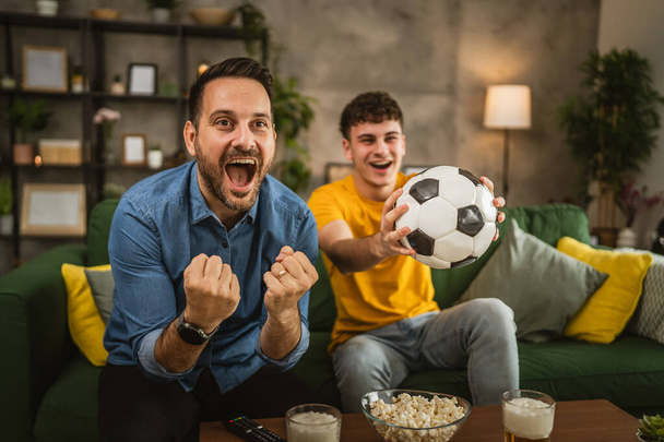 Young caucasian and adult caucasian man brothers or friends watch football and cheer match hold football ball with popcorn and beer on table at home - Fotografie, Obrázek