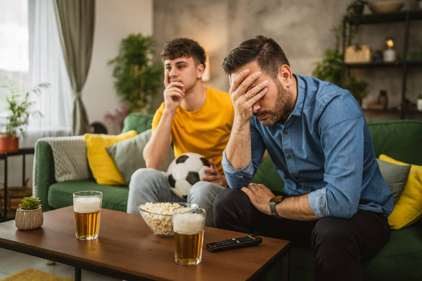 Dwóch mężczyzn przyjaciół kaukaskie są rozczarowani podczas oglądania meczu piłki nożnej, ponieważ ich zespół przegrywa mecz w domu - Zdjęcie, obraz