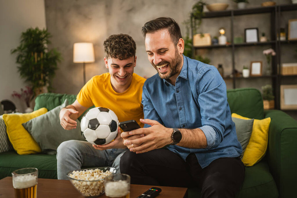 2人の男性の友人が自宅で携帯電話でサッカーの試合結果をフォローする - 写真・画像