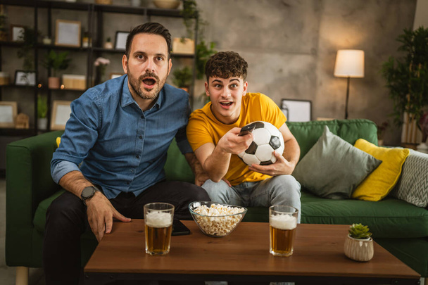 Νέοι καυκάσιοι και ενήλικες καυκάσιοι αδελφοί ή φίλοι βλέπουν ποδόσφαιρο και ευθυμία αγώνα κρατήσει μπάλα ποδοσφαίρου με ποπ κορν και μπύρα στο τραπέζι στο σπίτι - Φωτογραφία, εικόνα