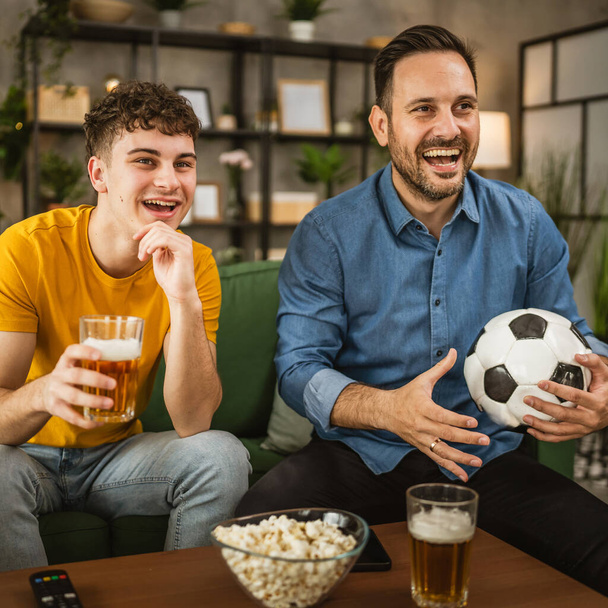 Joven caucásico y adulto caucásico hombre hermanos o amigos ver fútbol y animar partido celebrar pelota de fútbol con palomitas de maíz y cerveza en la mesa en casa - Foto, Imagen
