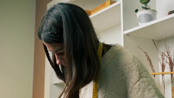 close-up em uma oficina de costura uma costureira no trabalho em uma camisola branca com centímetro amarelo em torno de seu pescoço - Filmagem, Vídeo