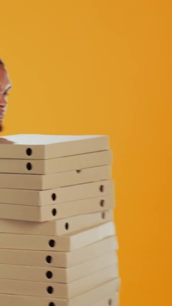 Dikey Video Erkek Pizza kuryesi stüdyoda devasa pizza kutuları taşıyor ve müşterilere yemek siparişi vermeye hazırlanıyor. Genç teslimatçı elinde koca bir paket hazır yemek, nakliye... - Video, Çekim