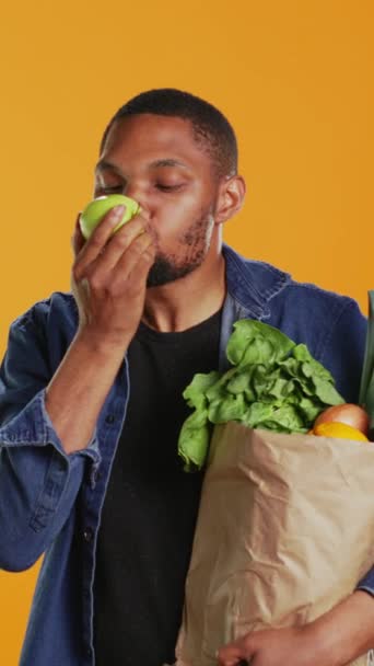 Vertical Video Afrikkalainen amerikkalainen vegaani kaveri puree kypsä luonnollinen vihreä omena, syö juuri korjattu orgaanisia hedelmiä studiossa. Paikallista maataloutta ja eettisesti tuotettuja tuotteita mainostava henkilö - Materiaali, video