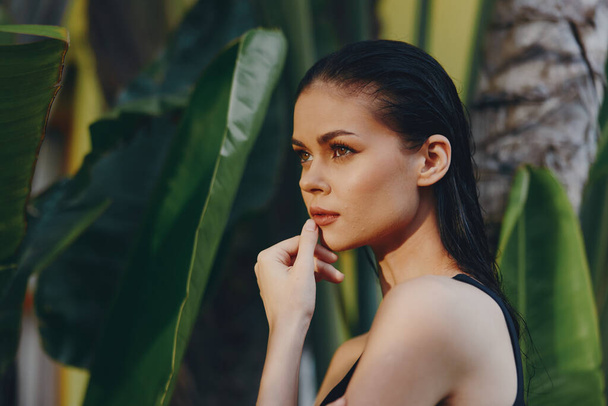 Portrait of a contemplative woman in a black tank top standing in front of a lush green plant, with hand on chin - Photo, Image