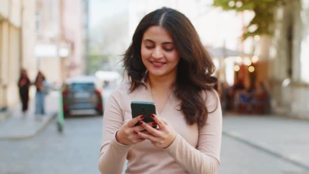 Ευτυχής χαμογελαστή νεαρή Ινδή γυναίκα που χρησιμοποιεί μηνύματα κειμένου smartphone περιήγηση στο διαδίκτυο, τελειώνοντας την εργασία, κοιτάζοντας κάμερα σε εξωτερικούς χώρους. Ισπανίδα που περπατάει στο δρόμο της πόλης. Τρόπος ζωής στην πόλη - Πλάνα, βίντεο