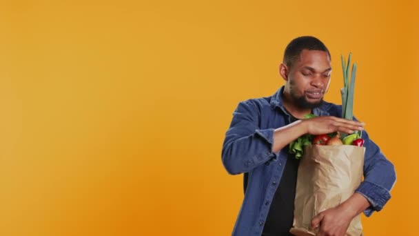 Счастливый молодой человек рекламирует органические фрукты и овощи местного производства, представляя биохимические химикаты бесплатно морковь в студии. Радостный человек поддерживает здоровое питание и веганство. Камера B. - Кадры, видео