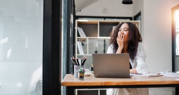 Une femme est assise à un bureau avec un ordinateur portable et une tasse de café. Elle bâille et elle est fatiguée. - Photo, image