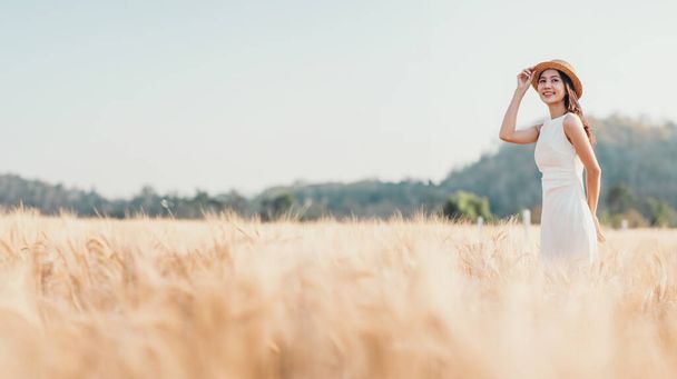Eine Frau steht in einem Feld mit hohen goldenen Weizen. Sie trägt ein weißes Kleid und einen Strohhut. Die Szene ist friedlich und heiter, die Frau blickt in die Ferne - Foto, Bild