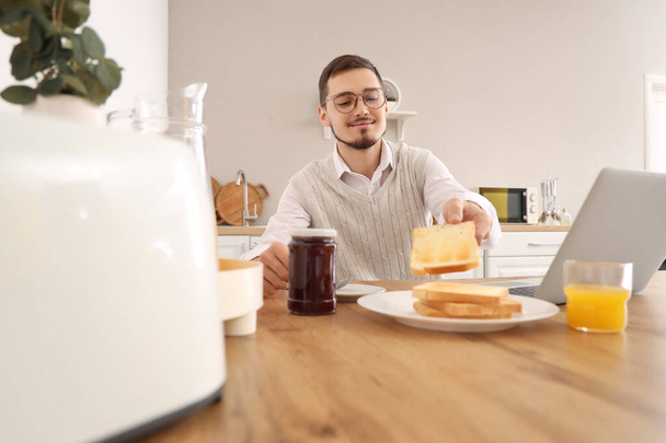 Fiatal férfi laptoppal, ízletes lekváros pirítós reggelire a konyhában - Fotó, kép