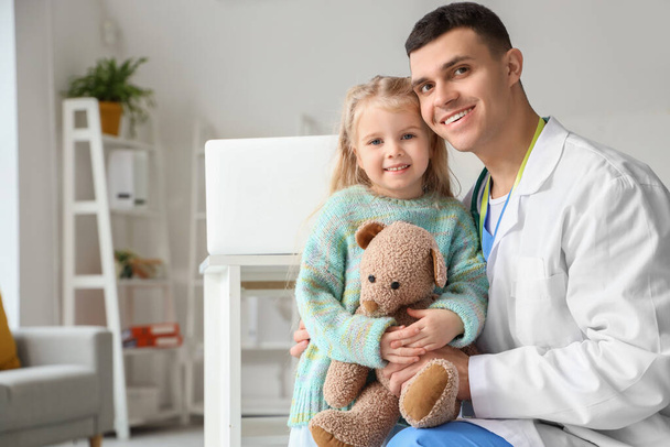 Male pediatrician and little girl with teddy bear in clinic - Фото, изображение