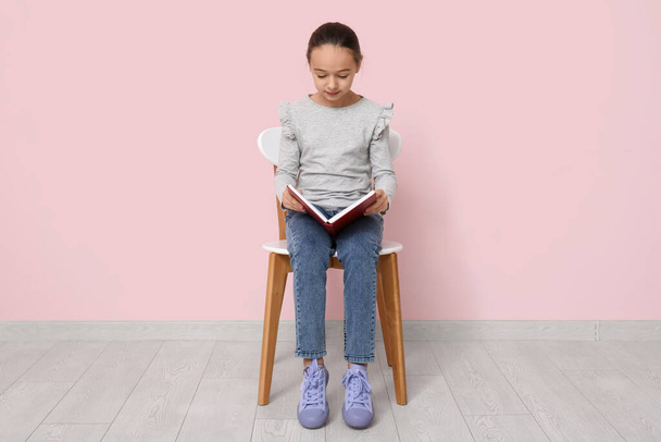 Petite fille lecture livre tout en étant assis sur la chaise près du mur rose - Photo, image