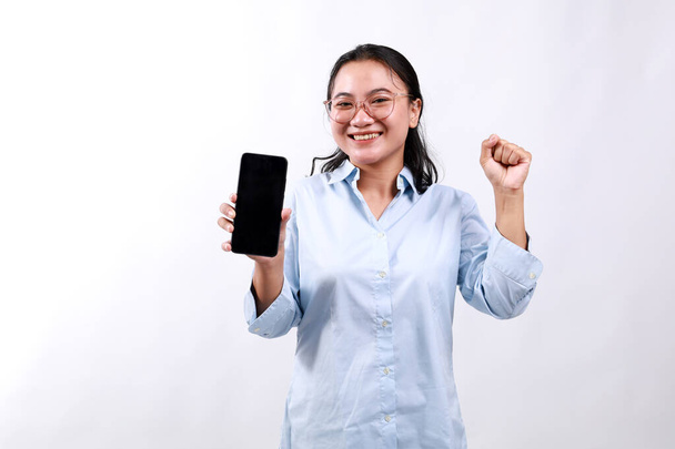 Ενθουσιασμένη νεαρή Ασιάτισσα, που δείχνει την εφαρμογή smartphone και θριαμβεύει, γιορτάζει στο κινητό τηλέφωνο, στέκεται πάνω από το λευκό φόντο - Φωτογραφία, εικόνα