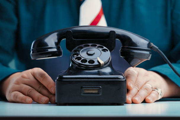 Hands rest beside an old-fashioned black rotary phone on a desk, the person wearing a teal jacket and a striped tie, suggesting readiness to communicate or provide assistance - Φωτογραφία, εικόνα