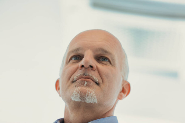 Vista de cerca de un hombre mayor con una expresión reflexiva, mirando hacia arriba, destacando su perilla blanca sobre un fondo brillante - Foto, imagen