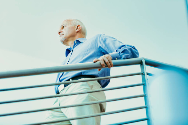 Старший человек в голубой рубашке прислоняется к перилам, вдумчиво глядя вдаль, размышляя о прошлом и будущем жизни - Фото, изображение