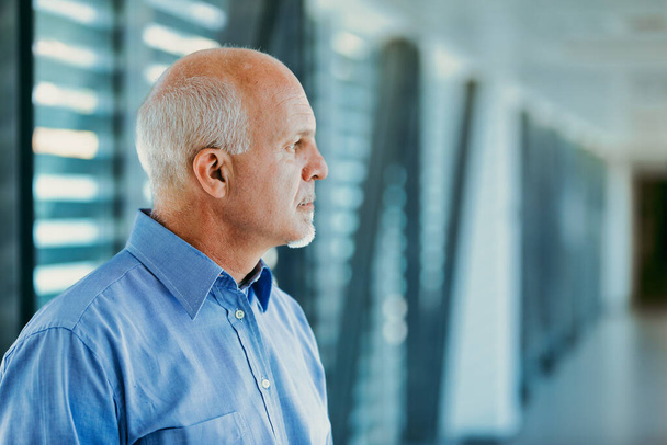 Αντανακλαστικός ηλικιωμένος άνδρας με φαλακρά λευκά μαλλιά και στοχαστική έκφραση φοράει μπλε πουκάμισο σε ένα θολό αρχιτεκτονικό περιβάλλον - Φωτογραφία, εικόνα
