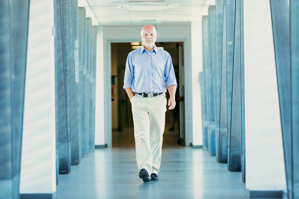 Hombre mayor con un paseo seguro, vestido con una camisa azul y pantalones ligeros, paseos en un pasillo bien iluminado - Foto, imagen