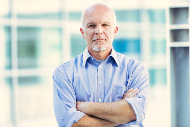 Senior professional stands confidently with arms crossed in front of a modern glass building, embodying seasoned leadership and expertise - Photo, Image