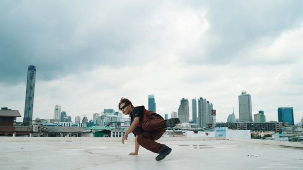 Съемка танцевального выступления би-боя профессиональной уличной танцовщицей на крыше с небоскрёбом, вид на город. Привлекательный азиатский хипстер показывает энергичный шаг. Открытый спорт 2024. Endeavor. - Фото, изображение