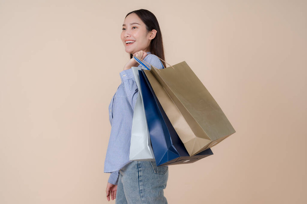 Joven mujer asiática alegre en ropa vaquera casual está sosteniendo múltiples bolsas de compras con una sonrisa radiante sobre un fondo pastel, expresando emoción y satisfacción después de ir de compras - Foto, imagen