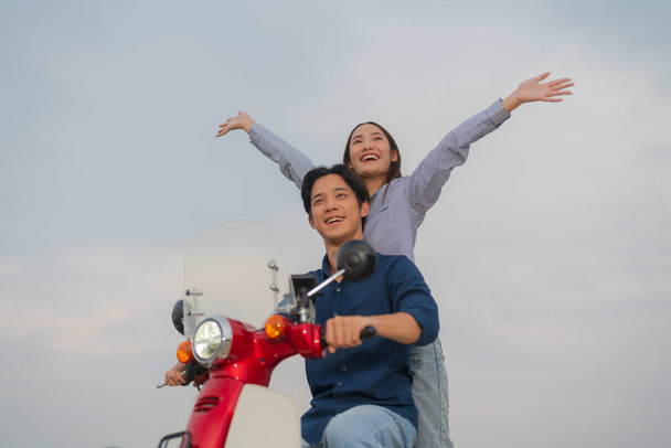 Νεαρό ζευγάρι ασιατών σε casual ρούχα απολαμβάνοντας μια διασκεδαστική βόλτα σε μια κόκκινη μοτοσικλέτα σε ένα ανοικτό δρόμο, με τη γυναίκα που υψώνει τα χέρια αισθάνονται ελεύθεροι και ευτυχισμένοι - Φωτογραφία, εικόνα